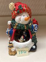 Frontal image of the porcelain snowman figurine showing the red and white hat with gold accents, green scarf, blue mittens, broom in hand, red cardinal perched on arm, and ice skating diorama visible in belly.