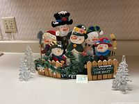 Wooden snowmen family behind a wooden picket fence decorated with greenery and pinecones, with a sign reading 'Snowmen warm your heart'. Three clear glass Christmas trees placed beside the wooden decoration.