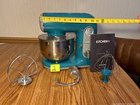 Front view of turquoise Kitchen HQ stand mixer with stainless steel bowl, flat beater, dough hook, wire whisk attachments, and manual. Tape measure shows approx 11 inches in width.