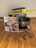 Sunbeam Mixmaster Deluxe mixer showing scale and included instruction and recipe manuals with attachments.