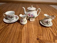 Porcelain tea set pieces displayed on wooden table showing teapot, two creamers, teacup and two saucers with floral designs.