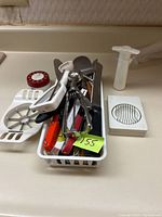 Photo of assortment of kitchen tools in a white plastic basket and items like apple cutter, pie lifter, peelers, cheese slicer and red timer visible on counter