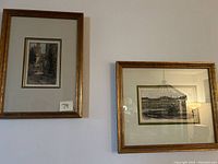 Photo of two framed etchings hanging on wall. Left frame has brownish-gold ribbed frame, etching shows narrow European street. Right frame has smooth gold frame, etching shows large palace building.