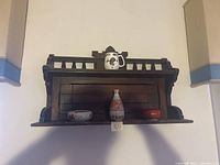 Overall view of dark wooden wall shelf with four displayed items: ceramic vase, ashtray, small red box, and mug with gold design.