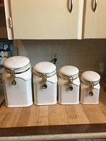 Four white airtight kitchen canisters in decreasing size order on wooden counter with tiled backsplash behind