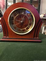 Front view of large wooden mantle clock showing Roman numeral dial, decorative hands, and brass-framed glass cover