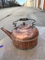 Copper pot placed outdoors showing side view with handle and spout, sitting on a protective surface.