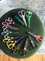 12 pairs of colorful crafting scissors arranged in a circle on a green round cushion, showing various decorative blade patterns and plastic handles.