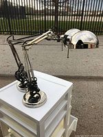 Two stainless steel adjustable desk lamps shown on a white table outside near a fence with the lamps in an upright position.