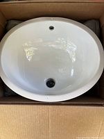 Top view of white oval ceramic sink with drain hole and overflow, inside shipping box.