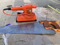 Photo showing Black & Decker electric sander with orange body and coiled cord alongside a saw with wooden handle and metal blade.