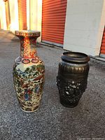Front view of two large vases placed side by side on pavement against a white wall with orange garage doors. Left vase is colorful porcelain, right vase is metallic dark with embossing.