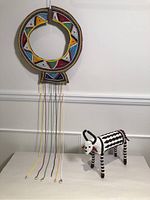 Image showing the full view of the circular beaded necklace with colorful geometric designs and long dangling strands alongside the black and white striped beaded water buffalo figurine.