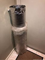 Roll of reflective foil foam backed insulation wrapped in plastic, standing upright on the floor against a wall.