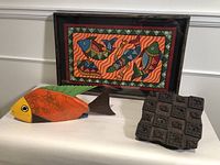 Frontal photo of three items on a white surface: framed painting propped upright, wooden fish sculpture lying flat, and wooden printing block stamp beside them.