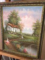 Full view of framed painting showing a cottage by a pond with two women in period dresses, floral foreground and green foliage, signed M. Jordan.
