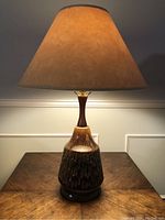 Full view of the mid century ceramic table lamp with a beige lampshade, ceramic body with textured brown glaze, wood neck, and base on wooden table.