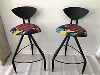 Photo showing two black metal frame bar stools with triangular black backrests and colorful geometric upholstered seats in Post Modern Memphis style.