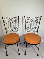 Pair of vintage bistro chairs with solid iron frames and orange vinyl round seats, showing front view with intricate scrollwork backs.