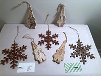 Set of five wooden holiday ornaments laid on table: two wood snowflakes with dark carved finish, three birch bark tree shapes, all with hanging cords.
