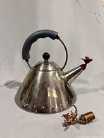 Front angled view of stainless steel kettle showing blue handle and red bird whistle on spout. Copper dragon whistle detached and lying beside, displaying the two whistles included.