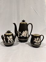 Full view of coffee pot, sugar bowl with lid, and creamer, all with Neo-classical white figures on black matte finish and gold accents.