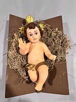 Front view of the painted plaster Christ Child statue with detailed facial features, glass eyes, and gold painted loincloth seated on the brown manger with straw moss bedding and gold metal halo.