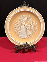 Front view of Franklin Mint 1979 Mother's Day plate with white relief of woman and girl on peach background on metal stand.