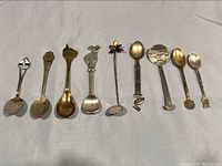 Overview photo showing all 9 pewter and Mexican silver souvenir spoons arranged side by side, displaying variety in handle motifs and shapes.