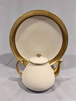 Front view of ivory teapot with gold accents placed on an ivory serving plate with a wide 24k gold border.