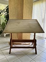 Front view of the drafting table with the top tilted upwards, showing the wood grain surface and sturdy wood frame base in ambient indoor lighting.