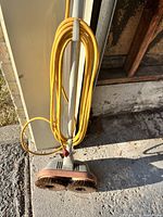 Floor polisher leaning against wall outside, showing dual circular brushes with yellow cord wrapped on handle.