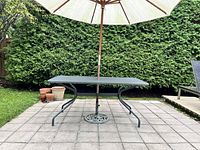 Full side view of rectangular outdoor patio table with beige patio umbrella inserted in umbrella hole. Table sits on gray square pavers with green bushes in background.