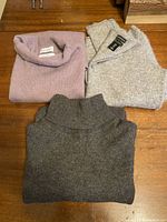 Three folded cashmere sweaters: muted lilac/pink turtleneck, charcoal grey turtleneck, and light grey round neck with buttoned neck opening, displayed on wood surface