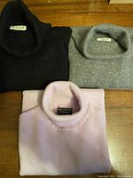 Three folded turtleneck sweaters in black, grey and pale pink/lilac displayed on a wooden surface.