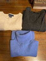 Three sweaters neatly folded on wooden surface: blue turtleneck, cream crewneck, grey cardigan