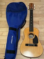 Full view of the Nova child's acoustic guitar next to the blue FirstAce soft guitar case on a hardwood floor.