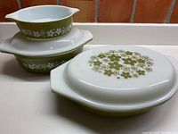 Overall view of three vintage Pyrex avocado green dishes with white Crazy Daisy floral pattern, stacked and arranged with lids visible.