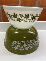 Photo showing two nesting bowls stacked, the smaller white bowl with green floral pattern inside the larger avocado green bowl with white floral pattern on their sides.