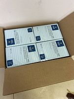Open cardboard box showing four stacks of sealed sticky note packs visible within. Each pack shows printed label with lines and size.