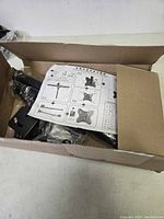 Open box showing black metal ceiling TV mount parts, installation screws, and paper instructions.