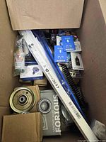 Top view of box showing assorted items including white LED strip light, metal pulley, various boxed parts, and a spray bottle