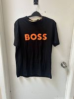Front view of black short-sleeve T-shirt with large orange BOSS logo across front, hanging on black hanger against white door.
