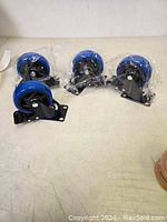 Four new black metal framed wheel castors with blue wheels arranged in a row, each wrapped in protective plastic.