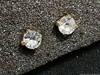 Close-up photo of a pair of small clear stone stud earrings with gold-tone prong settings on black textured background.