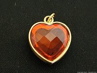 Close-up front view of a heart-shaped locket with gold finish and a large faceted amber-colored gem set in the center on a black background.