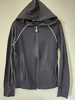 Front view of a black hoodie with full zipper, zippered pockets, and hood, showing white contrast stitching on shoulder seams