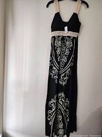 Front full view of black long dress with cream embroidery and lace straps hung on a hanger.