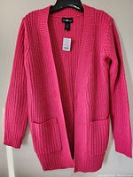 Full view of bright pink knit sweater jacket on hanger showing open front and two front pockets.
