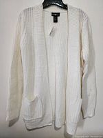 Full front view of ivory knit sweater jacket on black hanger showing open front, ribbed edges, and pockets.